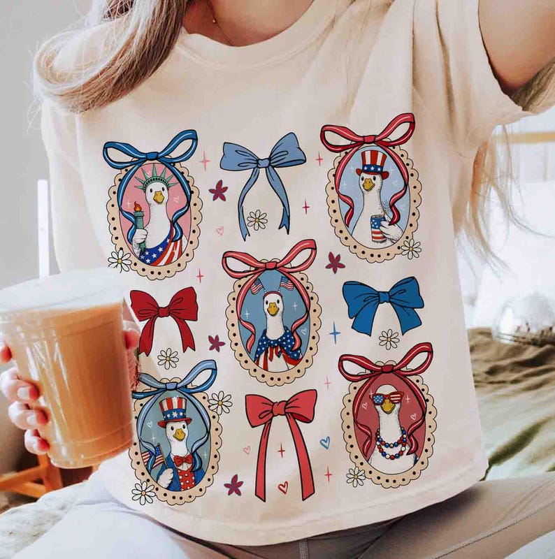 Op de afbeelding: Cr&egrave;mekleurig T-shirt met een grillig ontwerp van patriottische ganzen. Elke gans is omlijst in een decoratief ovaal, versierd met strikken en bloemmotieven. De ganzen zijn gekleed in rood, wit en blauw, met Amerikaanse vlaggen en vrijheidsfakkels.