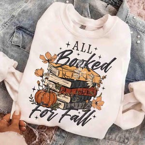 May include: White sweatshirt with a graphic design featuring a stack of books, a pumpkin, and floral accents. The text reads "All Booked for Fall" in a decorative font, perfect for autumn.