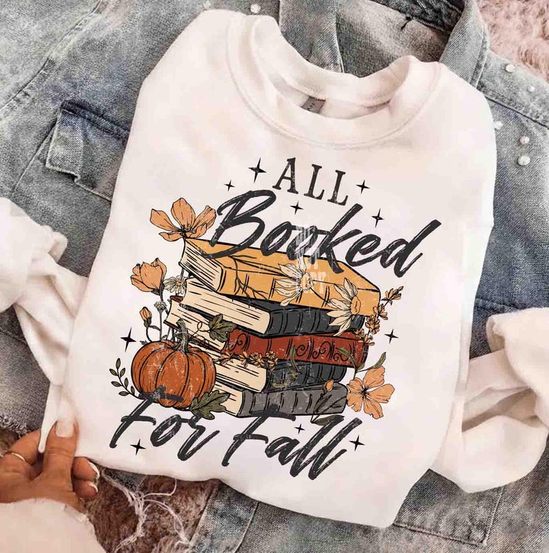May include: White sweatshirt with a graphic design featuring a stack of books, a pumpkin, and floral accents. The text reads "All Booked for Fall" in a decorative font, perfect for autumn.