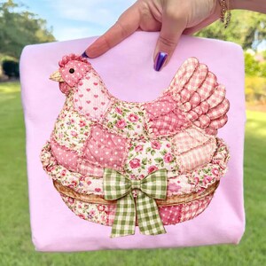 May include: A pink fabric chicken with a patchwork design, featuring hearts, floral patterns, and gingham. The chicken is set in a woven basket and has a green gingham bow. The background is a solid pink.