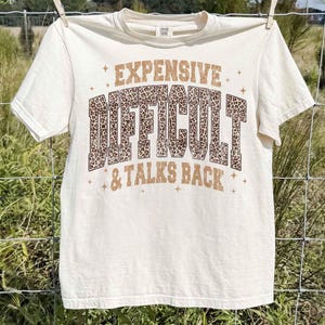 May include: Cream-coloured t-shirt with the words "EXPENSIVE, DIFFICULT, & TALKS BACK" in brown and leopard print lettering. The shirt is hanging on a clothesline.