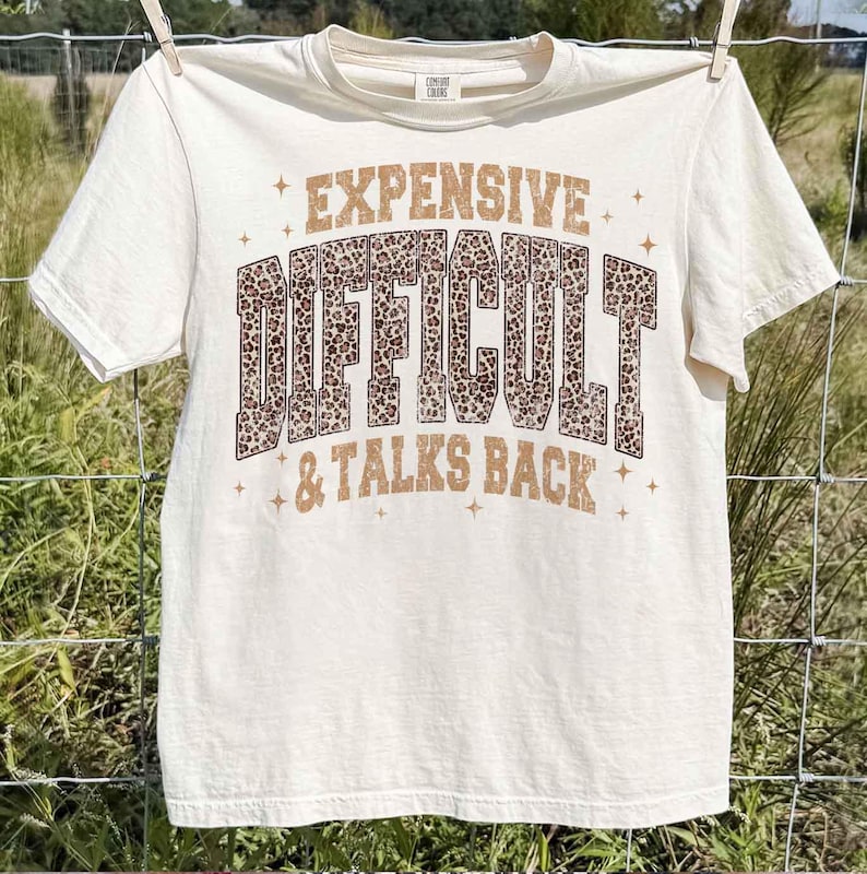 May include: Cream-coloured t-shirt with the words "EXPENSIVE, DIFFICULT, & TALKS BACK" in brown and leopard print lettering. The shirt is hanging on a clothesline.