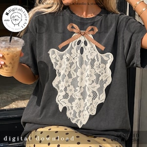 May include: A charcoal gray t-shirt featuring a large, white, lace-patterned ghost graphic centered on the chest. The ghost is adorned with a small, brown and white checkered ribbon bow at the top. The fabric has a faded, vintage-style appearance.