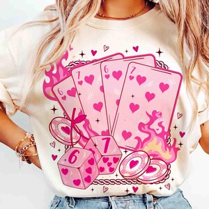 May include: Cream-colored sweatshirt featuring a pink graphic of playing cards, dice, and poker chips. The cards display hearts and the numbers 6 and 7. Pink flames and heart accents surround the design, creating a playful aesthetic.