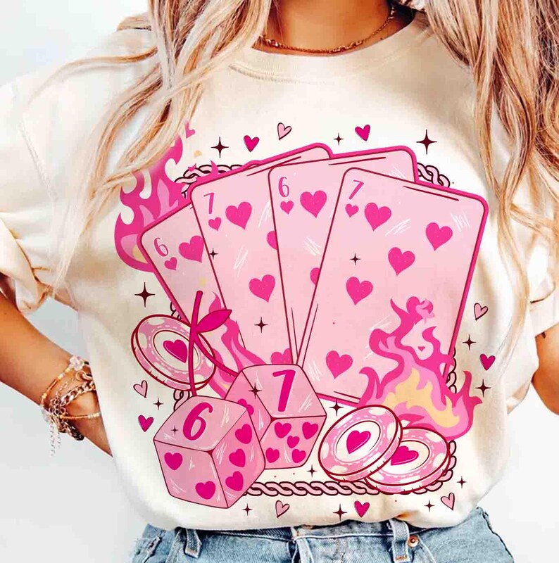 May include: Cream-colored sweatshirt featuring a pink graphic of playing cards, dice, and poker chips. The cards display hearts and the numbers 6 and 7. Pink flames and heart accents surround the design, creating a playful aesthetic.