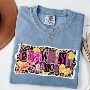 May include: A blue Comfort Colors t-shirt with the words "Crawfish Season" in a collage design. The design includes a crawfish, lemons, and corn on a leopard print background. A gold necklace is draped across the shirt.