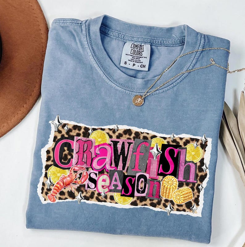 May include: A blue Comfort Colors t-shirt with the words "Crawfish Season" in a collage design. The design includes a crawfish, lemons, and corn on a leopard print background. A gold necklace is draped across the shirt.