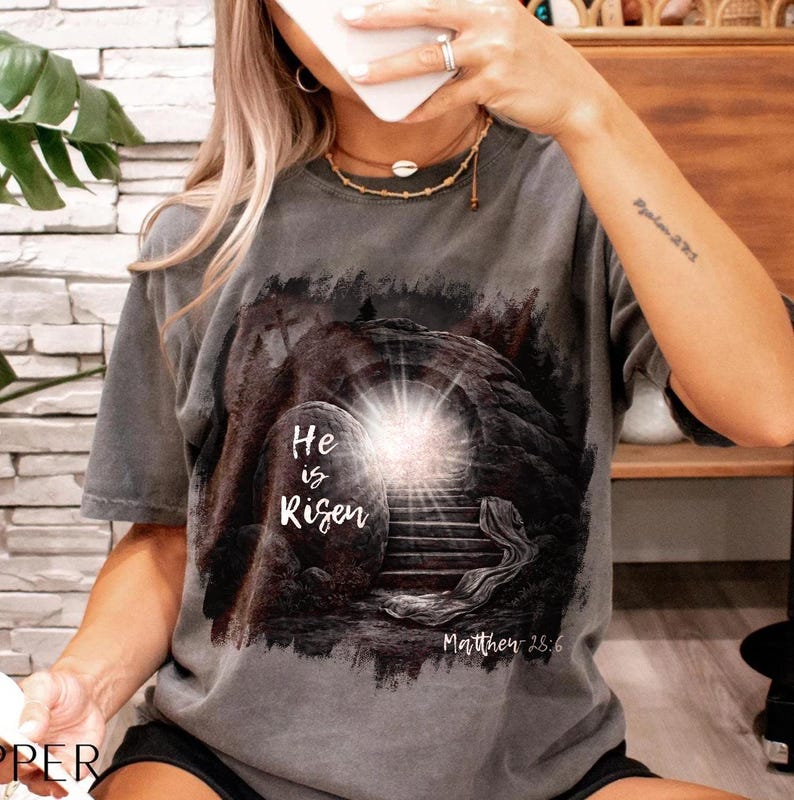 May include: Gray t-shirt featuring a graphic of an open tomb with a bright light emanating from within. The text "He is Risen" is displayed above the tomb, with "Matthew 28:6" below. The shirt has short sleeves and a crew neck.