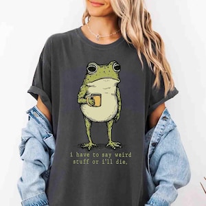 May include: Dark gray t-shirt featuring a cartoon frog holding a mug. The frog is green and beige with large eyes. The text "i have to say weird stuff or i'll die" is printed below the frog in a green font.
