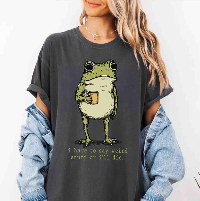 May include: Dark gray t-shirt featuring a cartoon frog holding a mug. The frog is green and beige with large eyes. The text "i have to say weird stuff or i'll die" is printed below the frog in a green font.