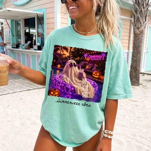 May include: A light teal t-shirt with a graphic of a ghost in sunglasses floating in a pool with pumpkins and the text "Summerween vibes". The ghost is holding a drink. The shirt is a relaxed fit.