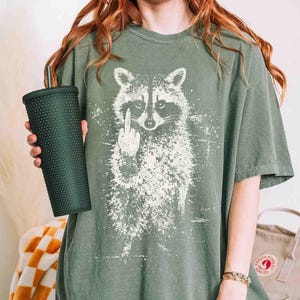 May include: Olive green t-shirt featuring a white graphic of a raccoon giving the middle finger. The shirt is worn with a dark green studded tumbler. The shirt has a vintage, distressed look.