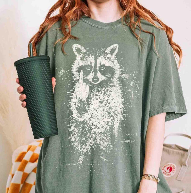 May include: Olive green t-shirt featuring a white graphic of a raccoon giving the middle finger. The shirt is worn with a dark green studded tumbler. The shirt has a vintage, distressed look.