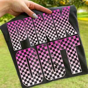 May include: Dark gray t-shirt with the words "PINK OUT" in large, distressed, pink and black checkered letters. The shirt is held up by a hand, with a blurred green background.