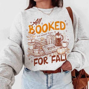 May include: A light gray sweatshirt with a fall-themed graphic. The design includes the text "All Booked For Fall" in orange and brown, along with illustrations of books, a pumpkin, coffee, and plants.