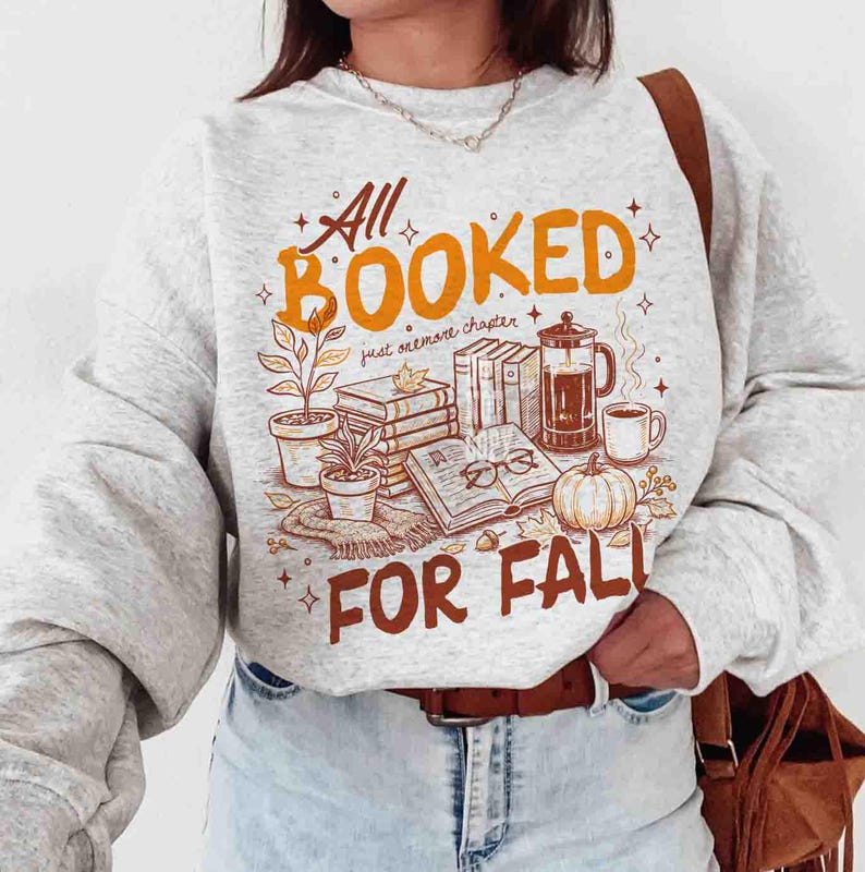 May include: A light gray sweatshirt with a fall-themed graphic. The design includes the text "All Booked For Fall" in orange and brown, along with illustrations of books, a pumpkin, coffee, and plants.