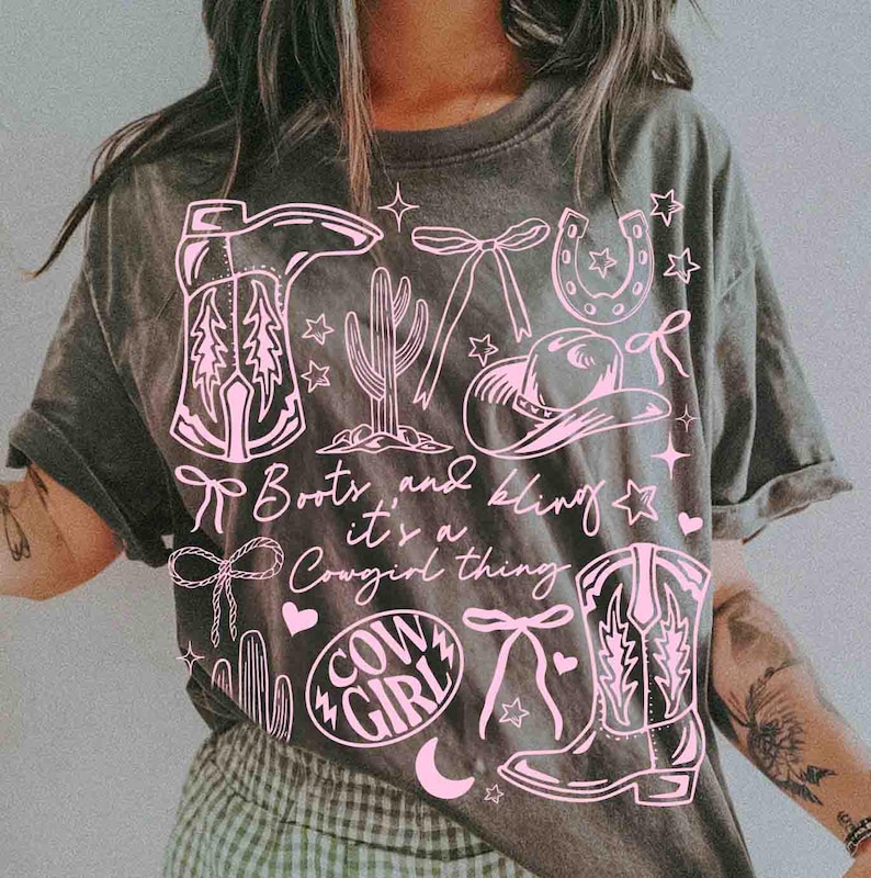 May include: Dark gray t-shirt with a pink graphic design featuring cowboy boots, a cactus, a horseshoe, a cowboy hat, bows, stars, and the words "Boots and Bling it's a Cowgirl thing" and "Cowgirl".