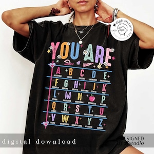 May include: A black t-shirt with the text "YOU ARE" in rainbow colors. The letters of the alphabet are listed with a positive adjective for each letter.  The design includes a variety of colorful illustrations, including hearts, stars, pencils, and an apple.