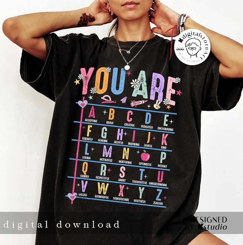 May include: A black t-shirt with the text "YOU ARE" in rainbow colors. The letters of the alphabet are listed with a positive adjective for each letter.  The design includes a variety of colorful illustrations, including hearts, stars, pencils, and an apple.