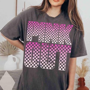 May include: Dark gray t-shirt with the words "PINK OUT" in a checkered pink and white design. The shirt has short sleeves and a relaxed fit, suitable for casual wear.