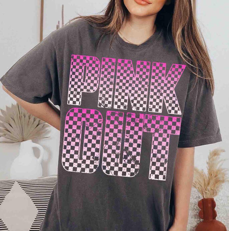 May include: Dark gray t-shirt with the words "PINK OUT" in a checkered pink and white design. The shirt has short sleeves and a relaxed fit, suitable for casual wear.