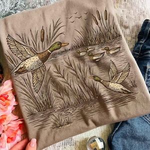 May include: A tan t-shirt featuring a detailed illustration of ducks in flight over a body of water. The design includes ducks, reeds, and flying birds, rendered in shades of brown and green. The shirt is paired with denim.