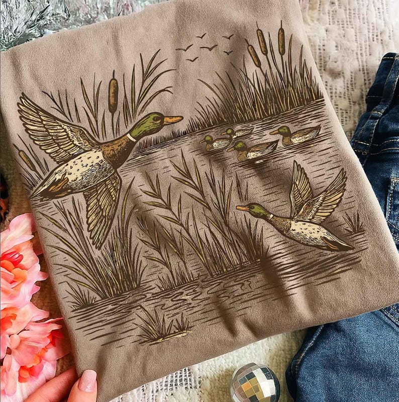 May include: A tan t-shirt featuring a detailed illustration of ducks in flight over a body of water. The design includes ducks, reeds, and flying birds, rendered in shades of brown and green. The shirt is paired with denim.
