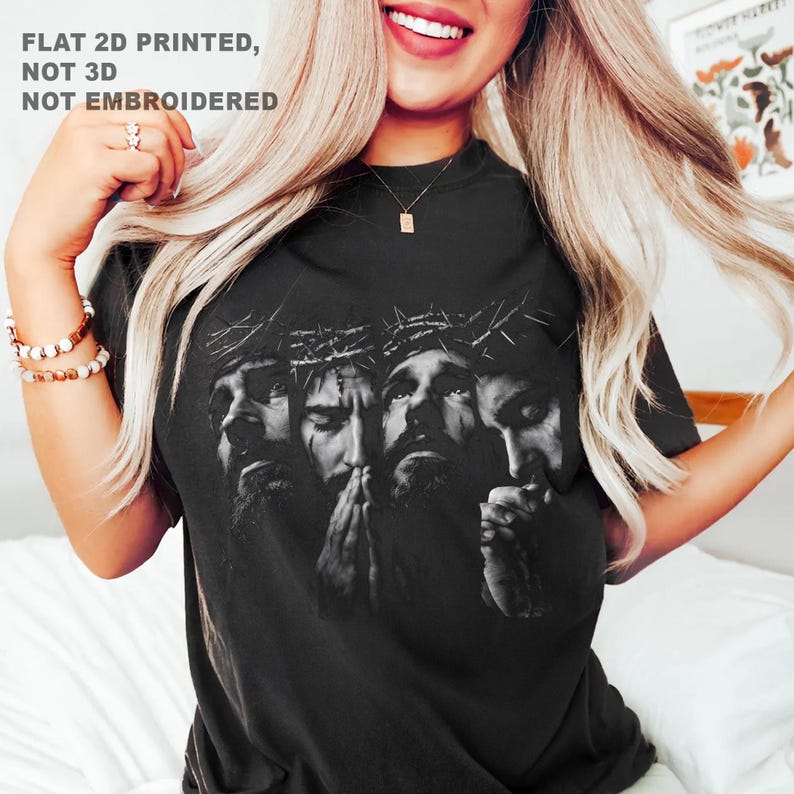 May include: Black t-shirt featuring a grayscale image of four faces, each wearing a crown of thorns. The text "FLAT 2D PRINTED, NOT 3D, NOT EMBROIDERED" is printed at the top. The shirt has a crew neck.