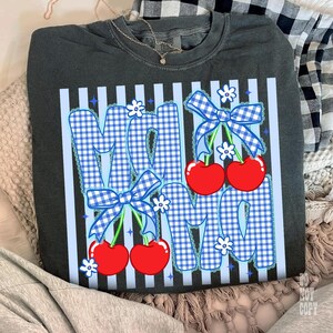 May include: Dark gray sweatshirt with a graphic design that says "MAMA" in blue gingham letters. The letters are decorated with blue bows, white flowers, and red cherries. The shirt has vertical light blue stripes.