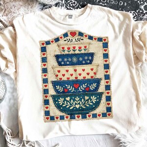 Pu&ograve; includere: T-shirt color crema con un design a ciotole impilate. Il design include ciotole blu e crema con cuori rossi e motivi floreali. Il design &egrave; incorniciato da un bordo blu e cuori rossi. La maglietta ha un'etichetta Comfort Colors.