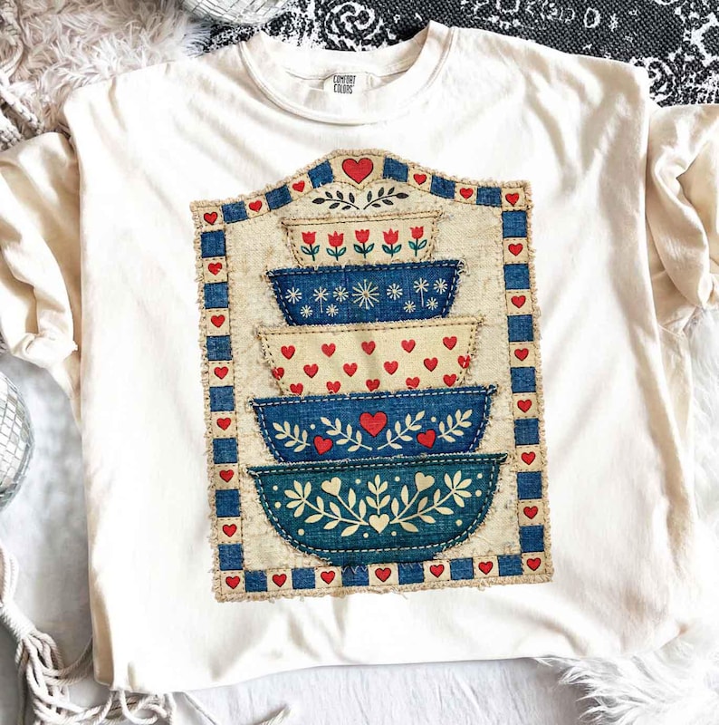 Pu&ograve; includere: T-shirt color crema con un design a ciotole impilate. Il design include ciotole blu e crema con cuori rossi e motivi floreali. Il design &egrave; incorniciato da un bordo blu e cuori rossi. La maglietta ha un'etichetta Comfort Colors.