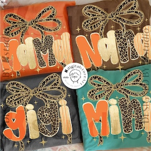 May include: Four colorful shirts with the words "Mama", "Nana", "Gigi", and "Mimi" in orange and gold lettering. Each shirt features a leopard print bow and star accents. The shirts are in shades of orange, brown, gray, and teal.
