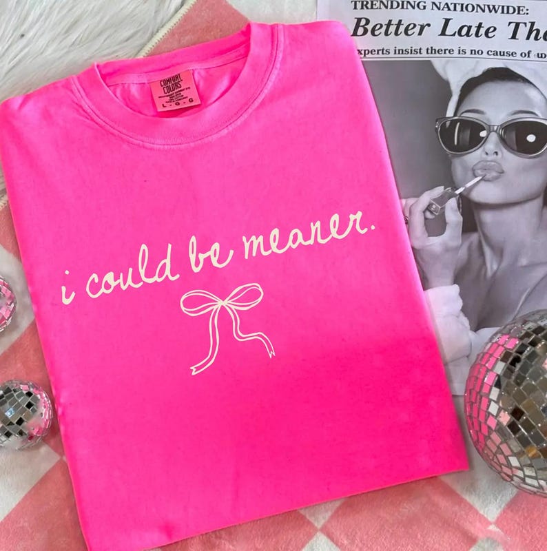 May include: A bright pink t-shirt with the phrase "i could be meaner" in white script and a white bow graphic. The shirt is folded on a pink and white checkered surface, with a black and white image in the background.