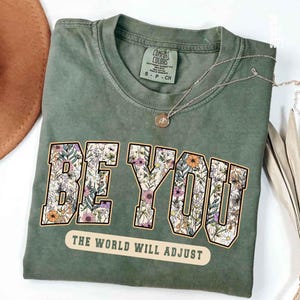 May include: Olive green t-shirt with the words "BE YOU" in a floral design. Below the text is the phrase "THE WORLD WILL ADJUST". The shirt has a crew neck and short sleeves. A silver necklace with a pendant is draped across the shirt.
