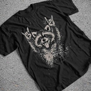 Pu&ograve; includere: T-shirt nero con una grafica invecchiata di un procione che fa il gesto rock and roll. Il procione &egrave; raffigurato in tonalit&agrave; di grigio e bianco, con la lingua di fuori. Il design ha un aspetto vintage e usurato.
