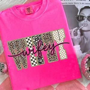 May include: Bright pink crew neck sweatshirt with the word "Wifey" in a bold, decorative font. The letters are filled with leopard print, black and white checkered patterns, and a tan and white checkered pattern. The word "wifey" is written in a cursive font.