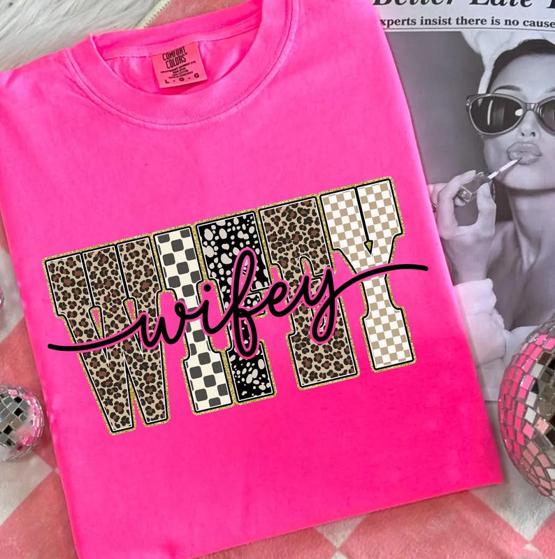 May include: Bright pink crew neck sweatshirt with the word "Wifey" in a bold, decorative font. The letters are filled with leopard print, black and white checkered patterns, and a tan and white checkered pattern. The word "wifey" is written in a cursive font.