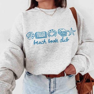 May include: A light gray sweatshirt with blue illustrations of a seashell, open book, crab, stacked books, and starfish. The words "beach book club" are written in blue script below the illustrations.