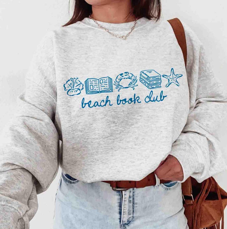 May include: A light gray sweatshirt with blue illustrations of a seashell, open book, crab, stacked books, and starfish. The words "beach book club" are written in blue script below the illustrations.