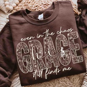 May include: A brown sweatshirt with the words "even in the chaos GRACE still finds me" in white script. The word "GRACE" is filled with a floral pattern. The sweatshirt is a crew neck style.