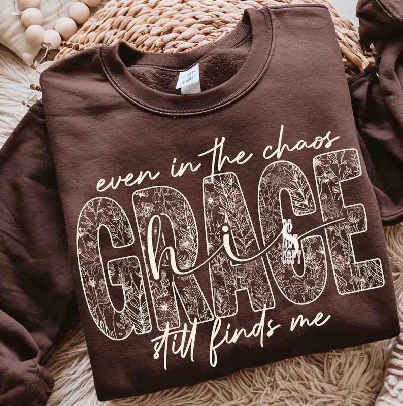 May include: A brown sweatshirt with the words "even in the chaos GRACE still finds me" in white script. The word "GRACE" is filled with a floral pattern. The sweatshirt is a crew neck style.