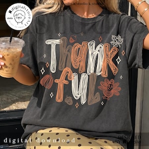 K&ouml;nnte beinhalten: Ein anthrazitfarbenes Kurzarm-T-Shirt mit dem Schriftzug Thankful in wei&szlig;em und orangefarbenem Linien-Design. Das Motiv enth&auml;lt Illustrationen eines Truthahns, von Eicheln, einem Ahornblatt und kleinen Rauten, die um den Text verteilt sind.
