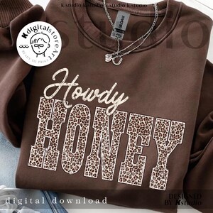 May include: A brown sweatshirt with the words "Howdy HONEY" in a white and leopard print design. The sweatshirt has a crew neck and long sleeves. A silver necklace is visible near the neckline. The words "digital download" are at the bottom.
