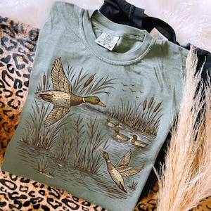 May include: Sage green Comfort Colors t-shirt featuring a detailed illustration of ducks in a marsh setting. The design includes flying ducks, reeds, and ducklings in the water. The shirt has a classic crew neck and a relaxed fit.
