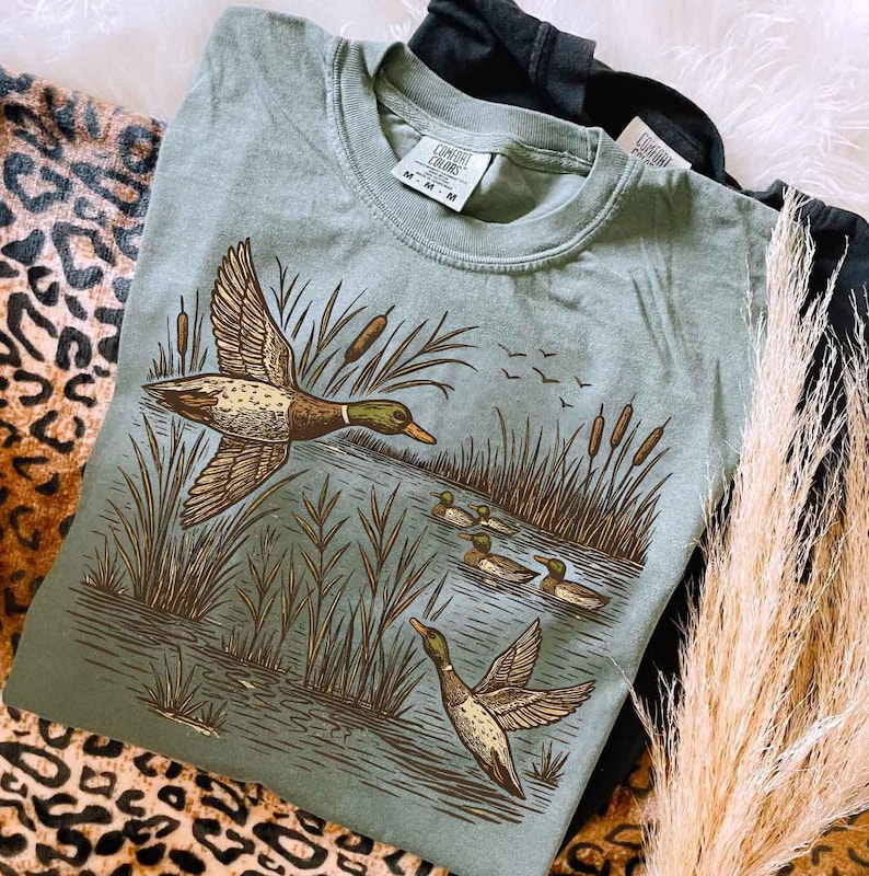 May include: Sage green Comfort Colors t-shirt featuring a detailed illustration of ducks in a marsh setting. The design includes flying ducks, reeds, and ducklings in the water. The shirt has a classic crew neck and a relaxed fit.
