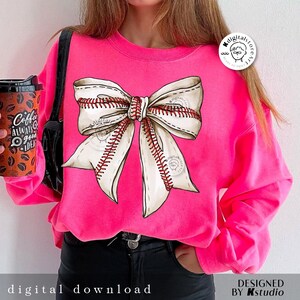 May include: A pink sweatshirt with a white and red baseball-themed bow on the front. The bow is made of white fabric with red stitching that resembles baseball seams.