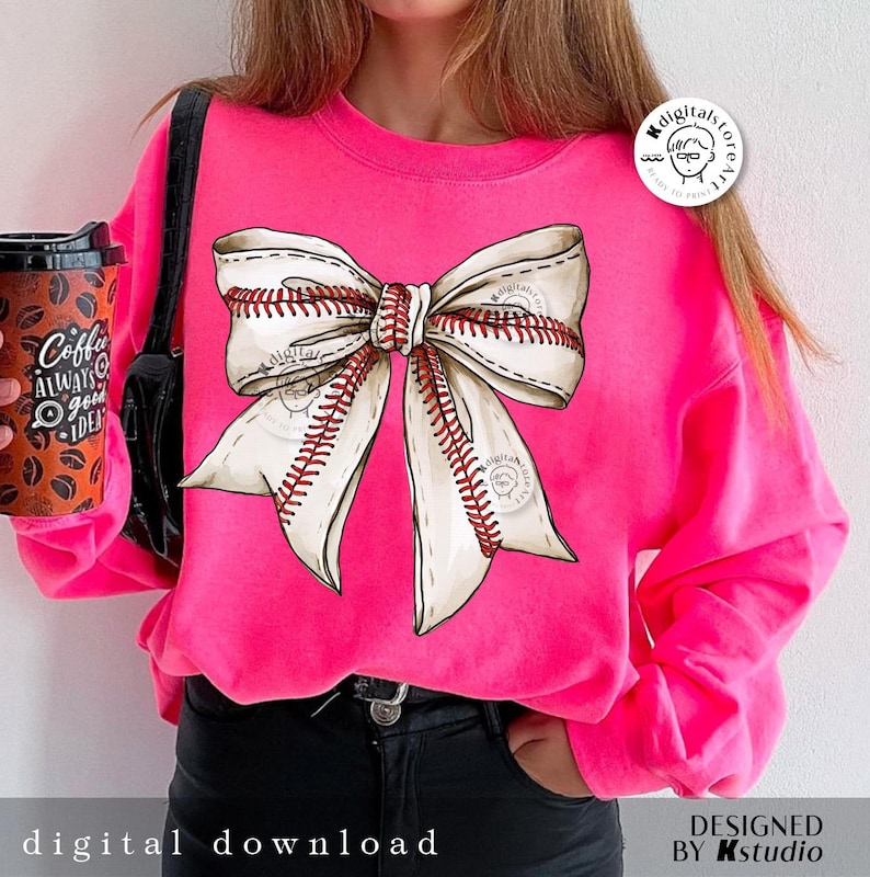 May include: A pink sweatshirt with a white and red baseball-themed bow on the front. The bow is made of white fabric with red stitching that resembles baseball seams.