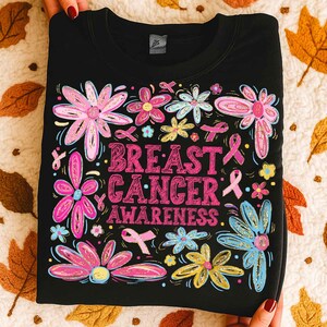 May include: Black sweatshirt with a floral design and the words "BREAST CANCER AWARENESS" in pink. The flowers are pink, blue, and yellow, with pink ribbons scattered throughout. The sweatshirt is folded and held by hands.