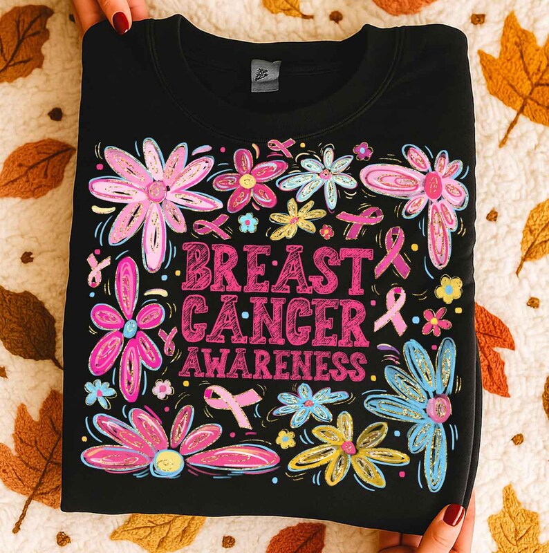 May include: Black sweatshirt with a floral design and the words "BREAST CANCER AWARENESS" in pink. The flowers are pink, blue, and yellow, with pink ribbons scattered throughout. The sweatshirt is folded and held by hands.