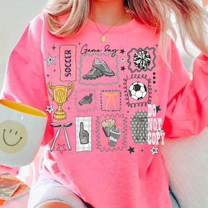 May include: A vibrant pink sweatshirt featuring soccer-themed graphics. The design includes a soccer ball, trophy, soccer boots, and the words "Game Day" and "Soccer". Other graphics include a pom-pom, a whistle, and a hand with the number one.